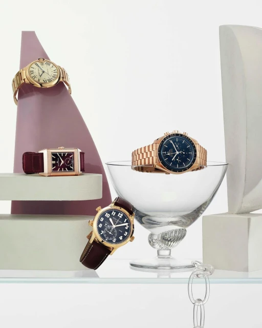 Still life arrangement featuring luxury wristwatches with gold and leather straps, displayed on geometric shapes and a glass bowl.