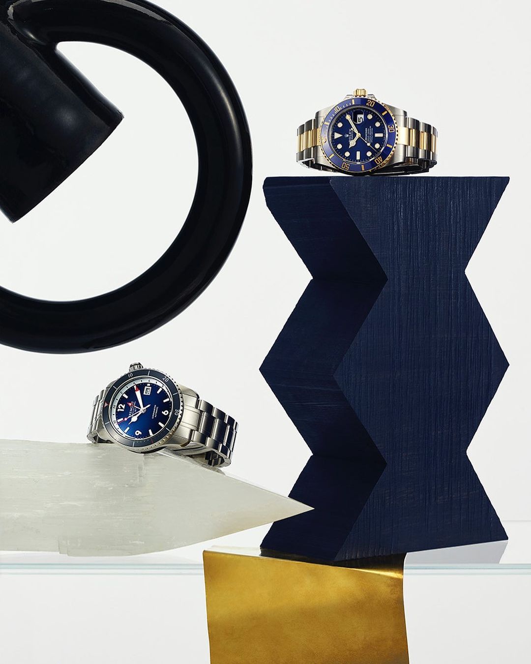 Two luxury wristwatches, one silver with a blue face and the other two-toned gold and silver with a blue face, on pedestals.