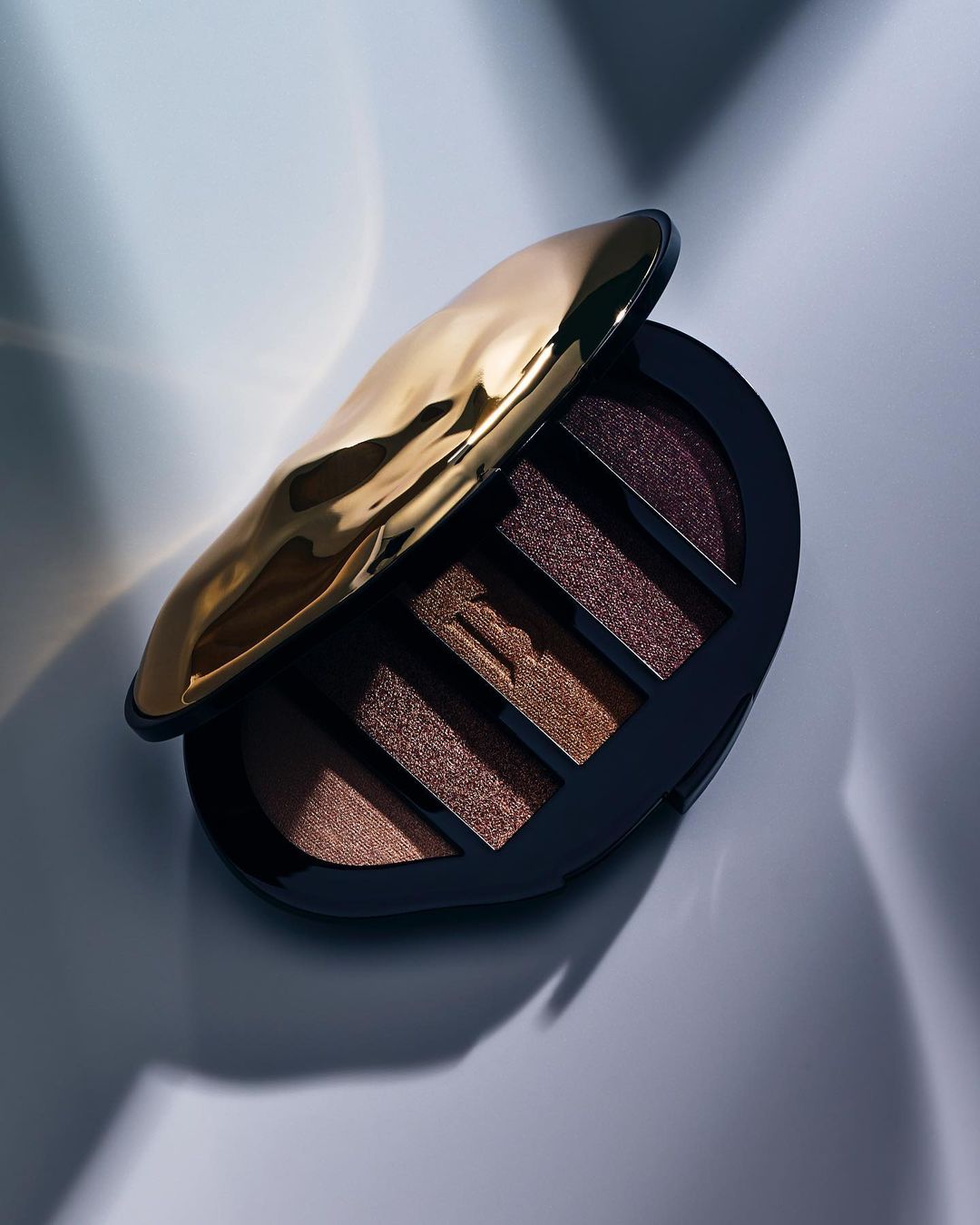 Elevated studio shot of an open eye shadow palette. Palette casing is black with a crinkled gold lid; shadows are earth-toned.