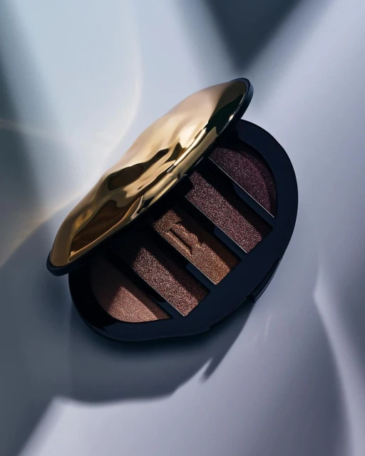 Elevated studio shot of an open eye shadow palette. Palette casing is black with a crinkled gold lid; shadows are earth-toned.