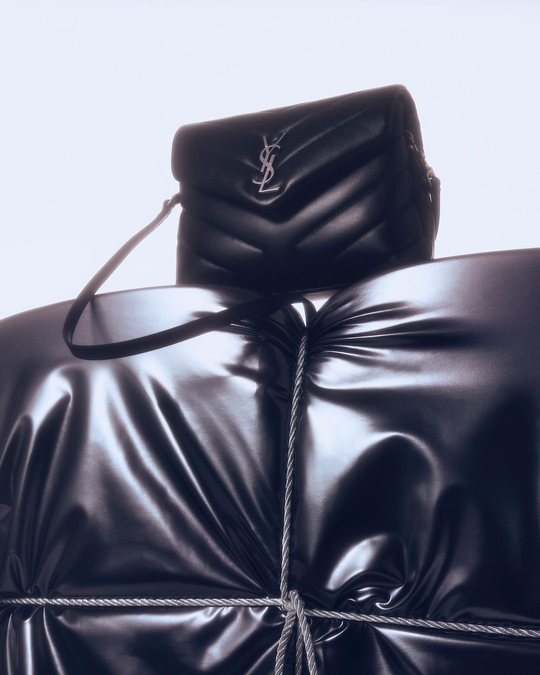 Black YSL shoulder bag on a glossy draped surface, possibly a garment. The studio product shot features a luxury brand against a bright background.
