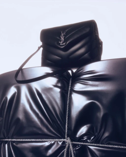 Black YSL shoulder bag on a glossy draped surface, possibly a garment. The studio product shot features a luxury brand against a bright background.