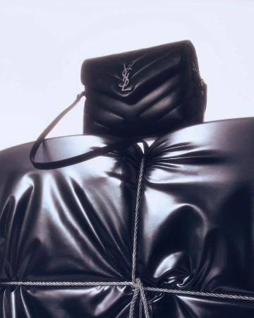 Black YSL shoulder bag on a glossy draped surface, possibly a garment. The studio product shot features a luxury brand against a bright background.