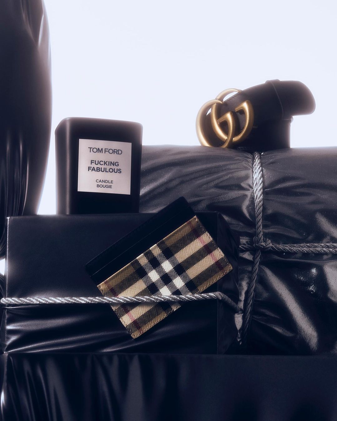 Still life featuring luxury branded accessories like a black Gucci belt, Burberry wallet, and Tom Ford candle on wrapped packages.