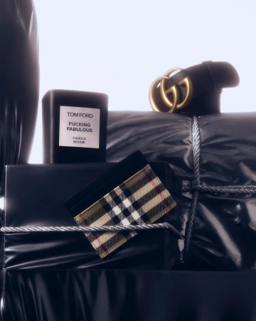 Still life featuring luxury branded accessories like a black Gucci belt, Burberry wallet, and Tom Ford candle on wrapped packages.