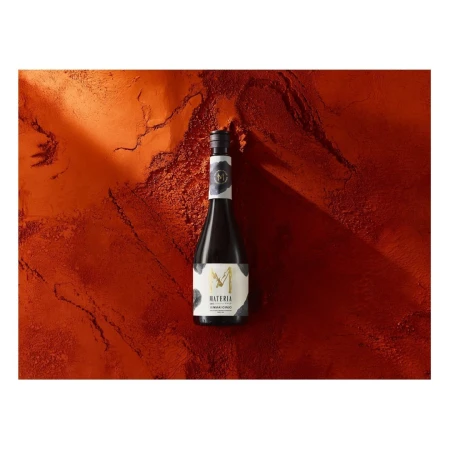 A dark glass bottle of Materia brand beverage resting vertically on a textured, rust-orange and terracotta-colored surface.