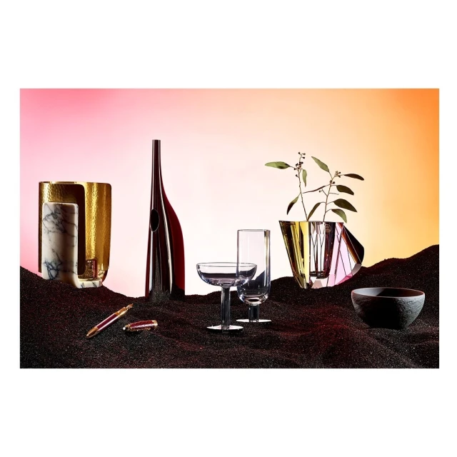 Still life photography showcases gold and marble vases, glass bottles, crystal glasses, a stone bowl, and pens on a bed of black granules.