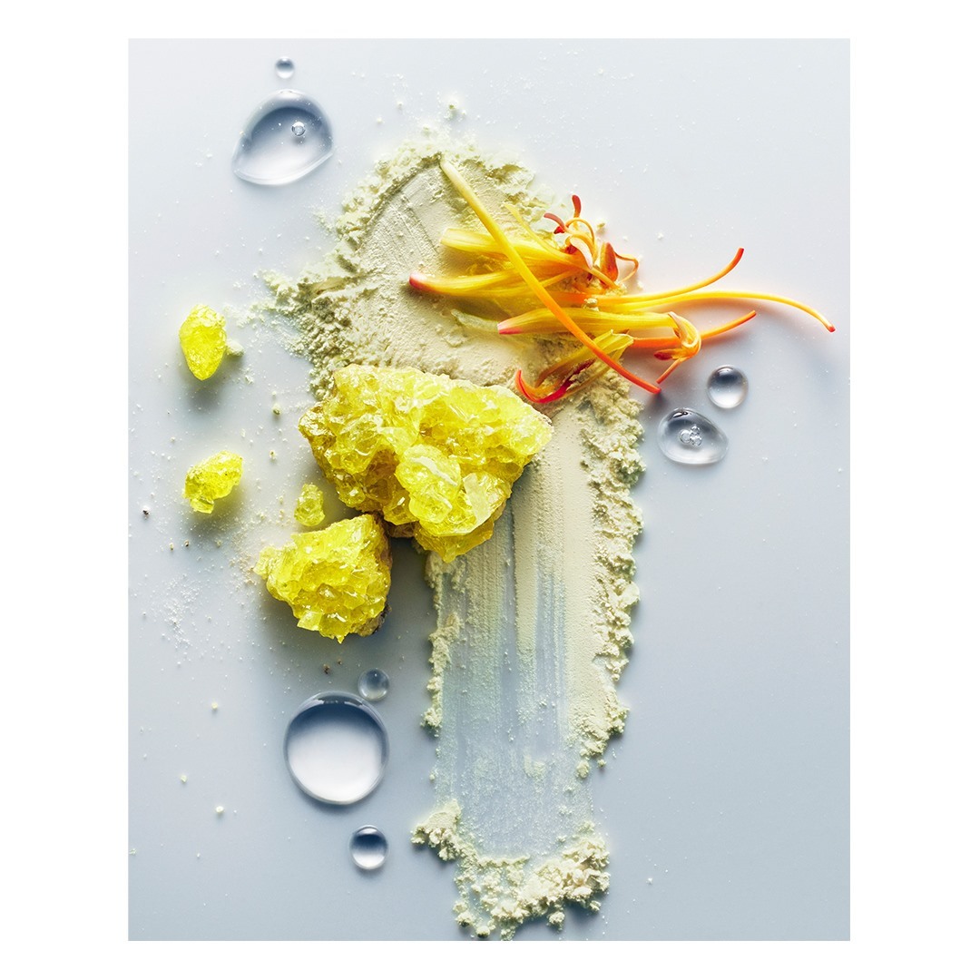 Macro studio shot of yellow crystals, clear water droplets, and powder with orange floral accents against a clean, bright white background.