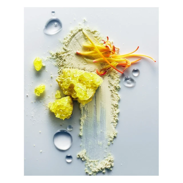 Macro studio shot of yellow crystals, clear water droplets, and powder with orange floral accents against a clean, bright white background.