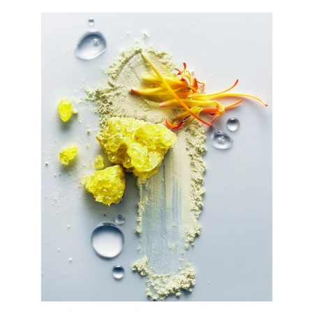 Macro studio shot of yellow crystals, clear water droplets, and powder with orange floral accents against a clean, bright white background.