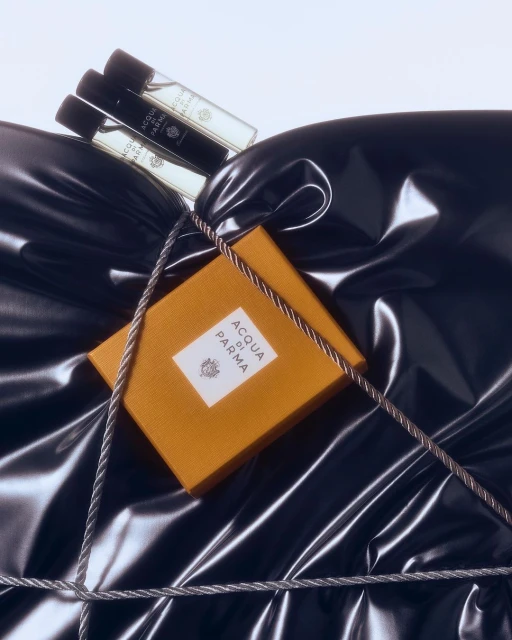 Close-up of Acqua di Parma cologne bottles and a yellow box on a glossy black fabric surface, tied with gray string, product photography.