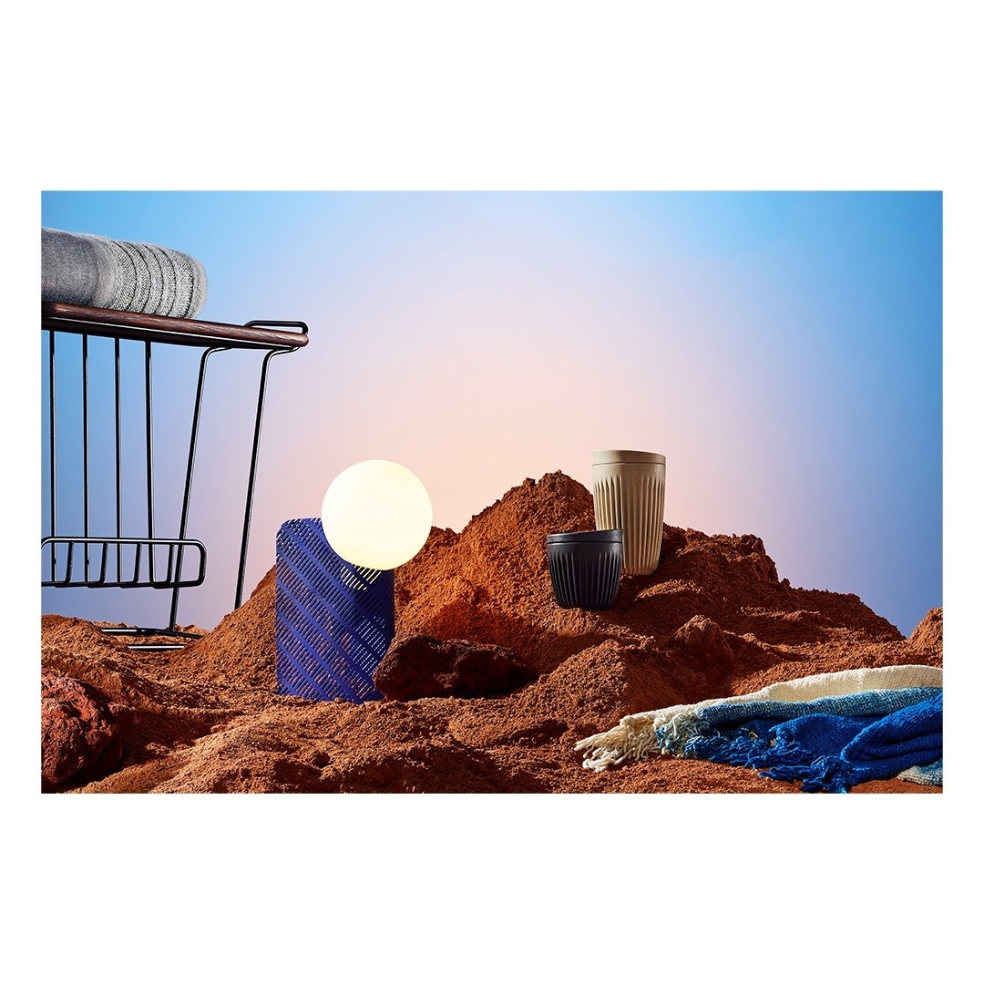Still life of household objects, including drinking cups, set against a gradient sky background and artificial desert sand surface.