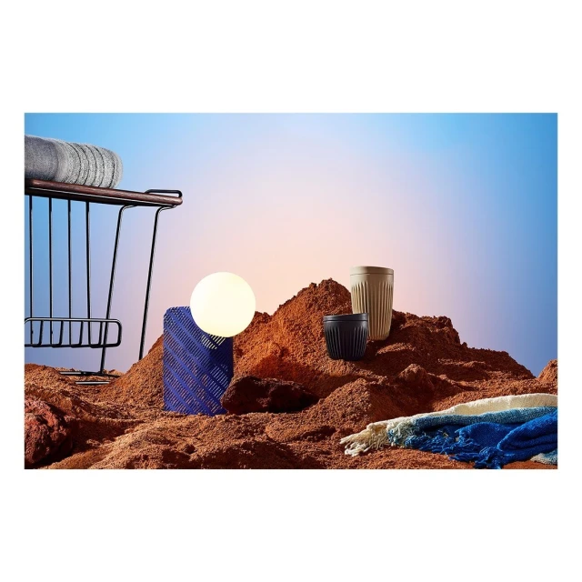 Still life of household objects, including drinking cups, set against a gradient sky background and artificial desert sand surface.