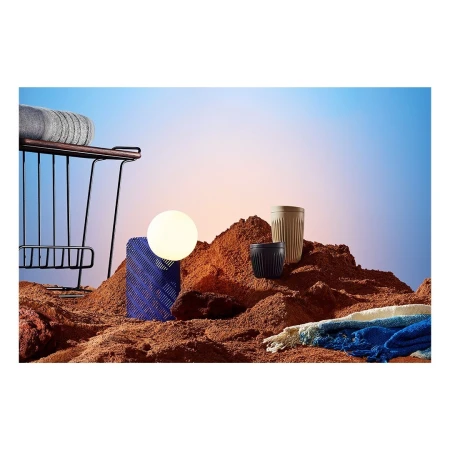 Still life of household objects, including drinking cups, set against a gradient sky background and artificial desert sand surface.