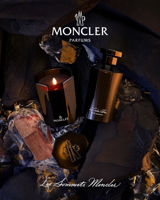 Moncler fragrance and candle on dark textural surface with logo. Perfume bottle and candle present brand in product shot.