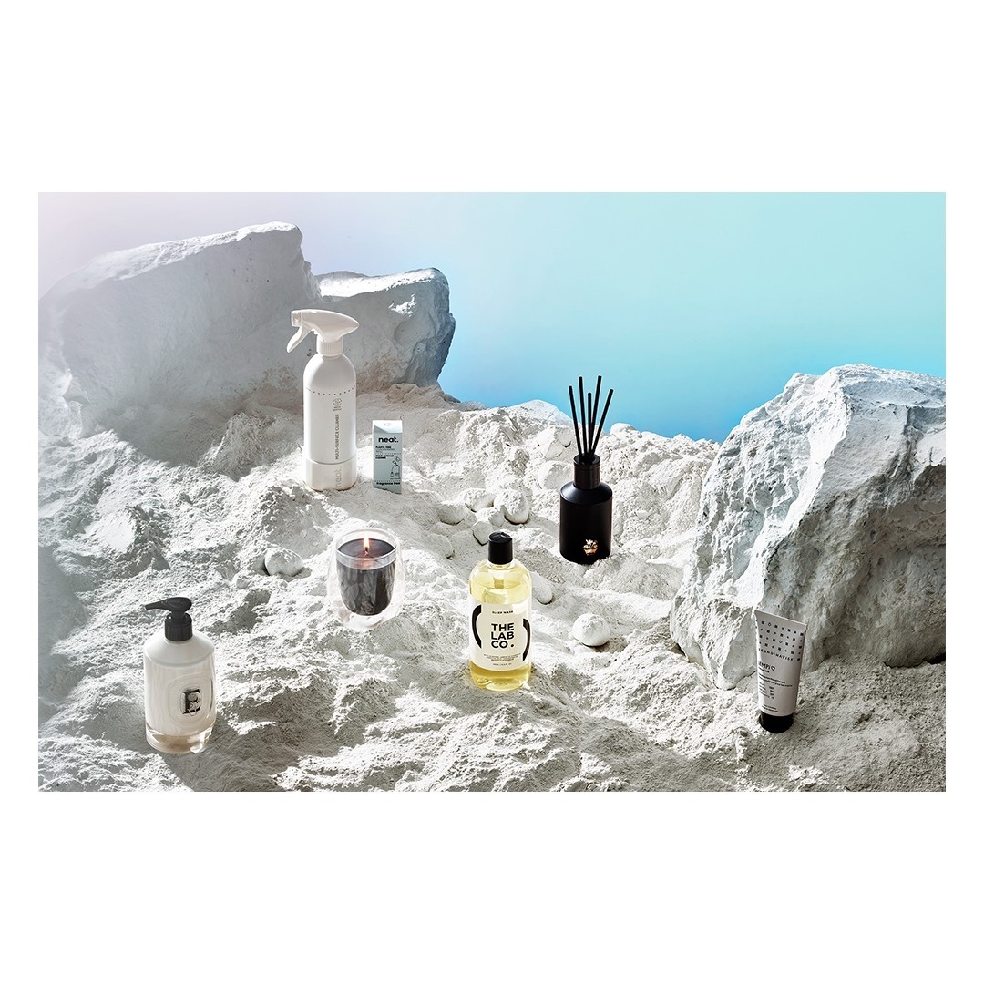 Still life of skincare and home fragrance products including lotion, cleanser, spray, candle, and diffuser on a textured white surface.