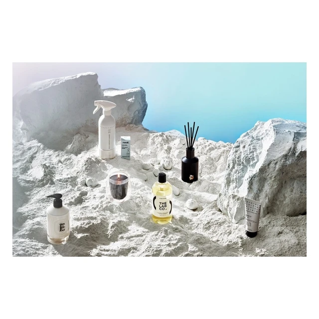 Still life of skincare and home fragrance products including lotion, cleanser, spray, candle, and diffuser on a textured white surface.