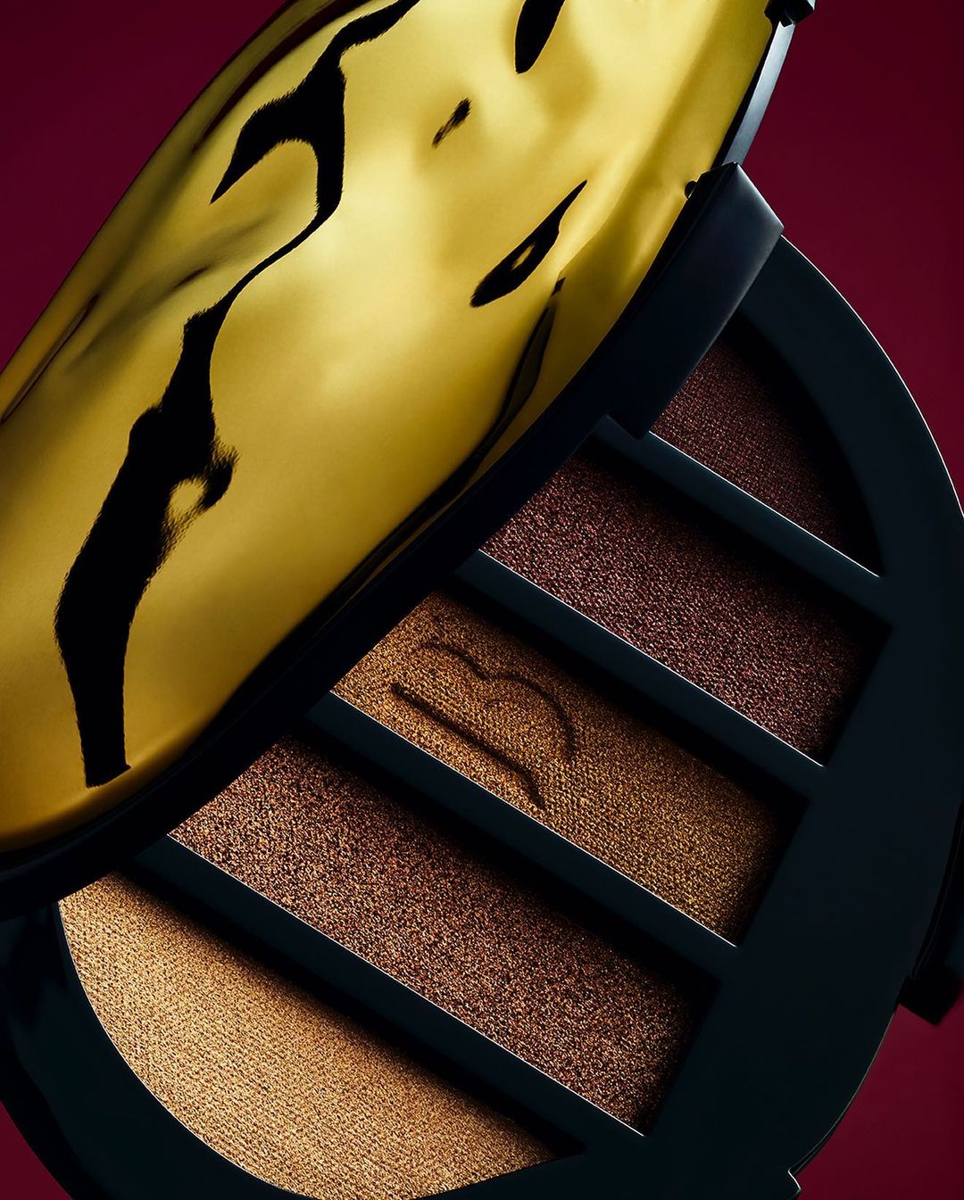 Close-up of a luxurious eyeshadow palette with gold and brown shades in a black case, presented against a rich burgundy background.