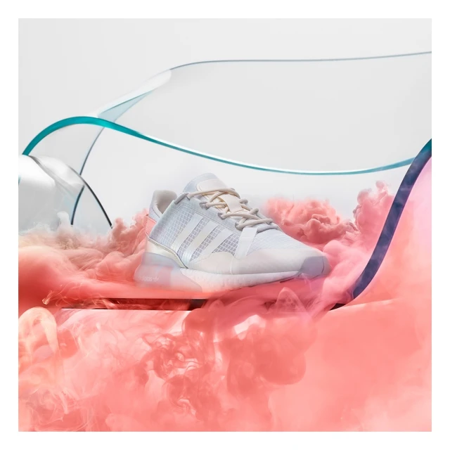 Promotional shot of a white Adidas sneaker surrounded by pink smoke, against a white backdrop and translucent blue accents, in studio.