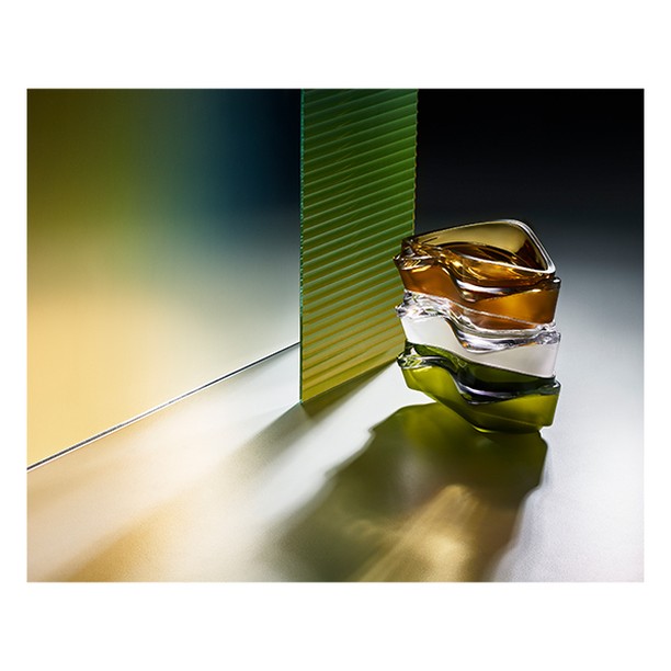 Abstract still life featuring stacked sculptural glass vases in yellow, white, and green, set against a background of textured glass.