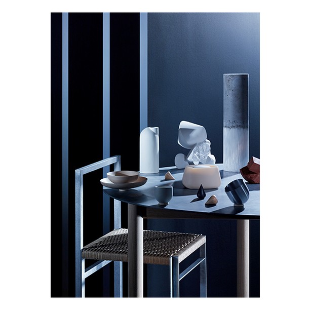 Still life featuring minimalist vases, bowls, and geometric shapes on a table with a metal chair against a dark blue and black background.
