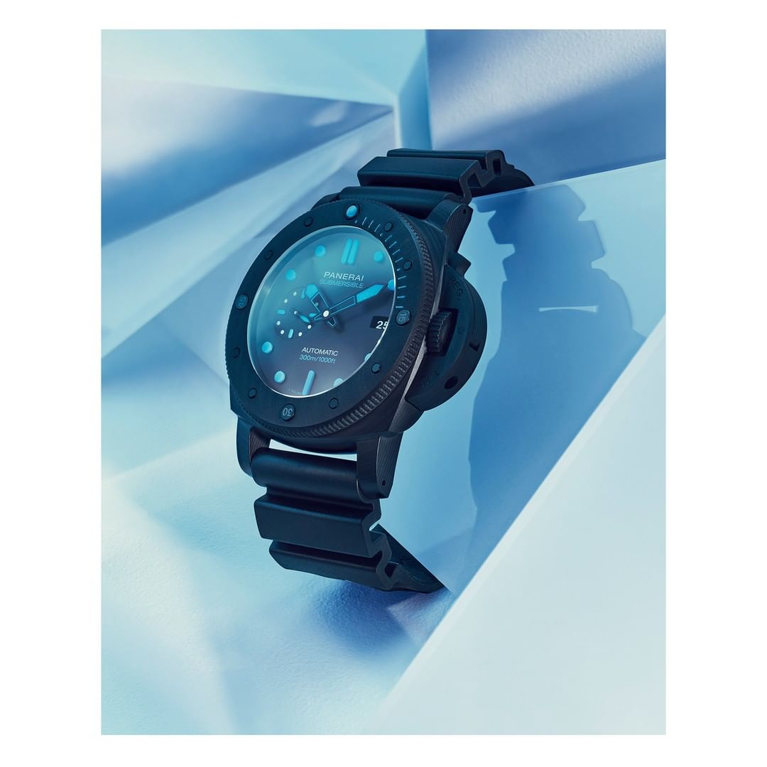 Black Panerai Luminor Marina watch on a blue geometric surface, showcasing its dial, bezel, and rubber strap in a studio product shot.