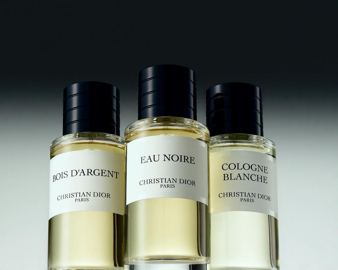 Three Christian Dior cologne bottles are lined up against a gradient gray background, featuring the brand's Eau Noire and Bois D'Argent scents.