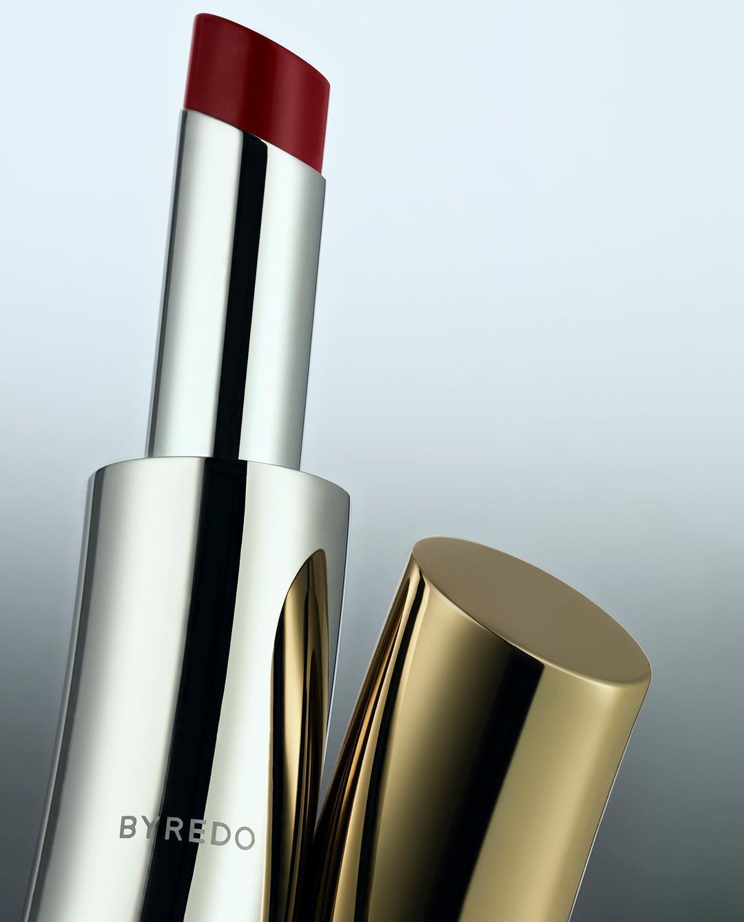 Close-up shot of red lipstick with a metallic silver and gold container against a gradient grey background, showcasing luxury cosmetics.