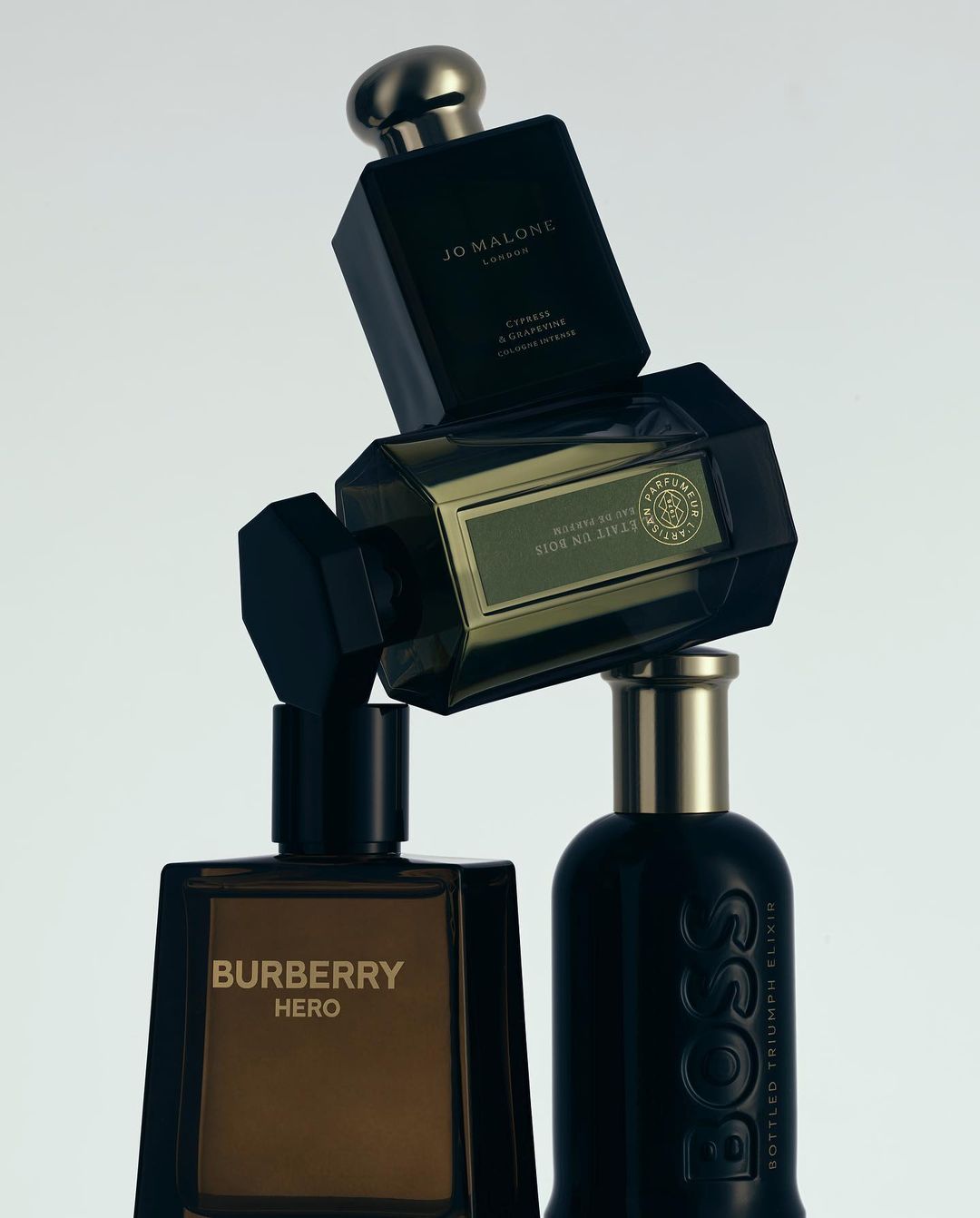 Four perfume bottles, including Burberry Hero, Jo Malone London, and Hugo Boss, stacked in front of a plain white studio background.