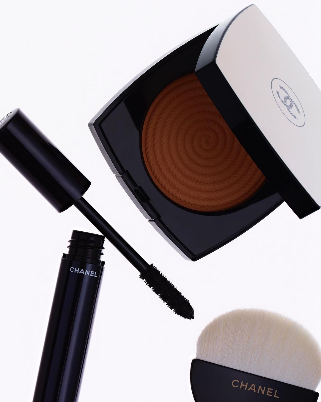 Chanel bronzer, mascara, and makeup brush are displayed against a white backdrop, highlighting the luxury beauty product arrangement.