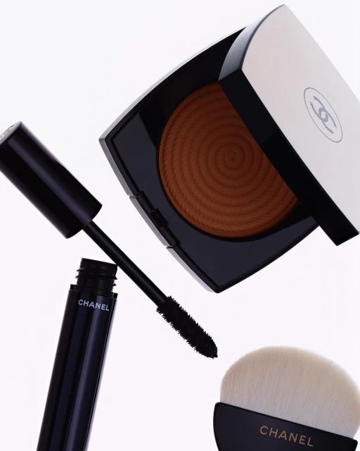 Chanel bronzer, mascara, and makeup brush are displayed against a white backdrop, highlighting the luxury beauty product arrangement.