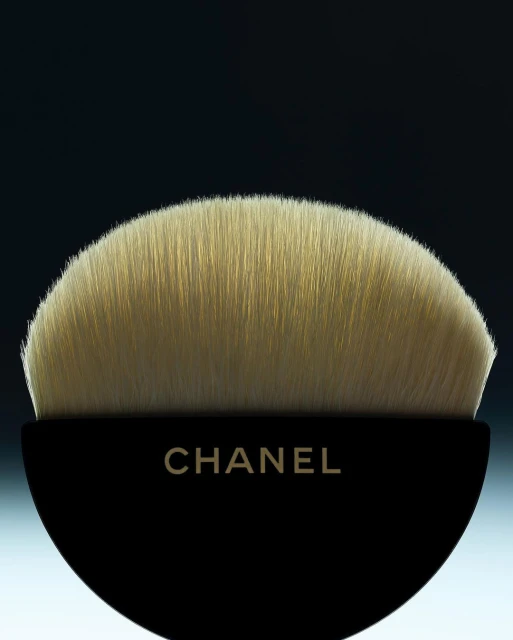 Close-up of a Chanel makeup brush with soft, tan bristles and a sleek, black handle against a soft, gradient background.