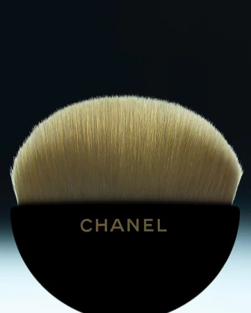 Close-up of a Chanel makeup brush with soft, tan bristles and a sleek, black handle against a soft, gradient background.