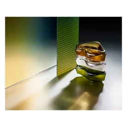 Stacked amber, clear, and green glassware sculptures on a grey surface with a fluted green panel and gradient backdrop