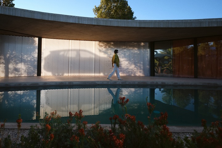 Adult walking near a reflecting pool with a modern building, lattice-patterned walls, and hints of greenery on a sunny day.