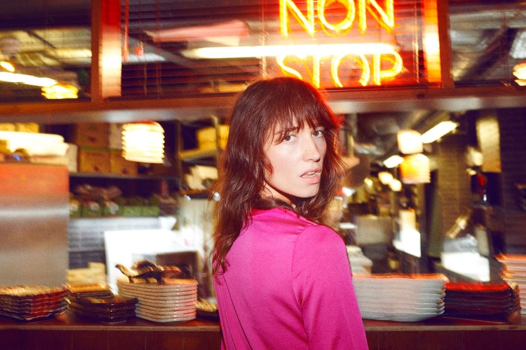 Woman in pink top stands in restaurant interior with a neon sign reading NON STOP, focusing on her face and fashion.