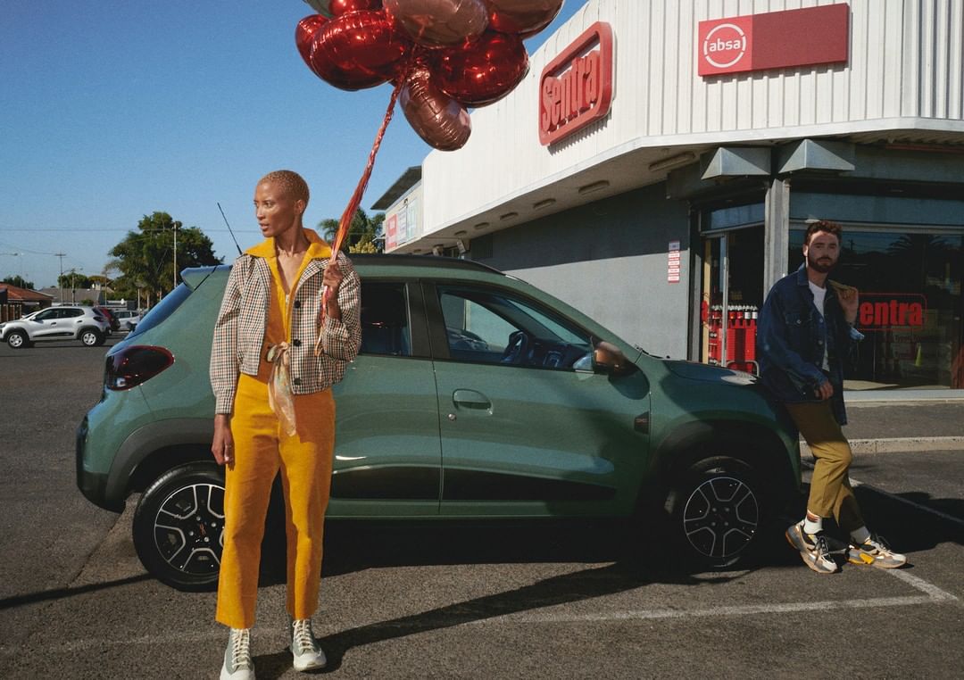 A green electric car is parked outdoors with a man and woman and red heart balloons in front of a Sentra grocery store under a blue sky.