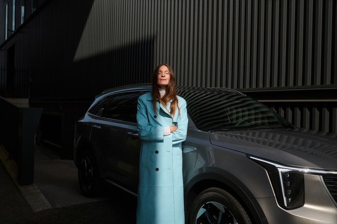 Adult female model in a blue coat standing in front of a grey SUV car with a dark wall in the background during the day.