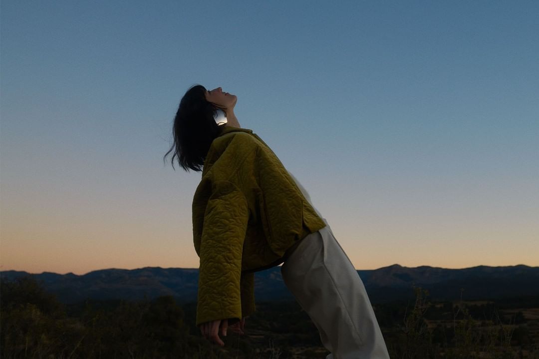 Woman in a yellow jacket tilts her head back towards the blue sky in a scenic outdoor landscape at dusk, evoking a sense of freedom.