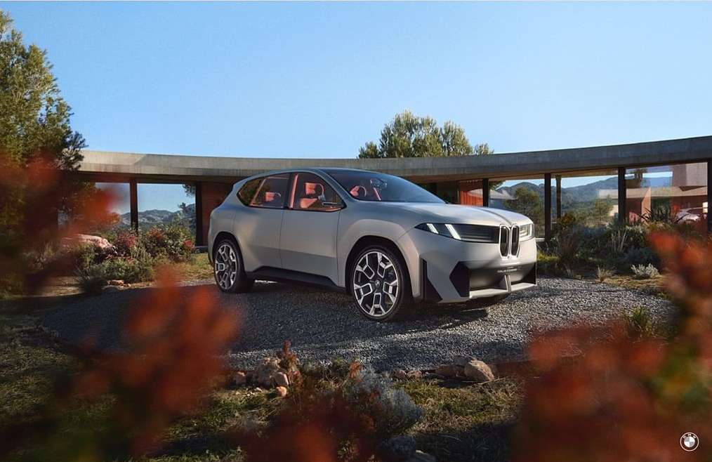 Silver BMW SUV parked on a gravel surface outside a modern home with large windows, under a clear sky, surrounded by greenery.
