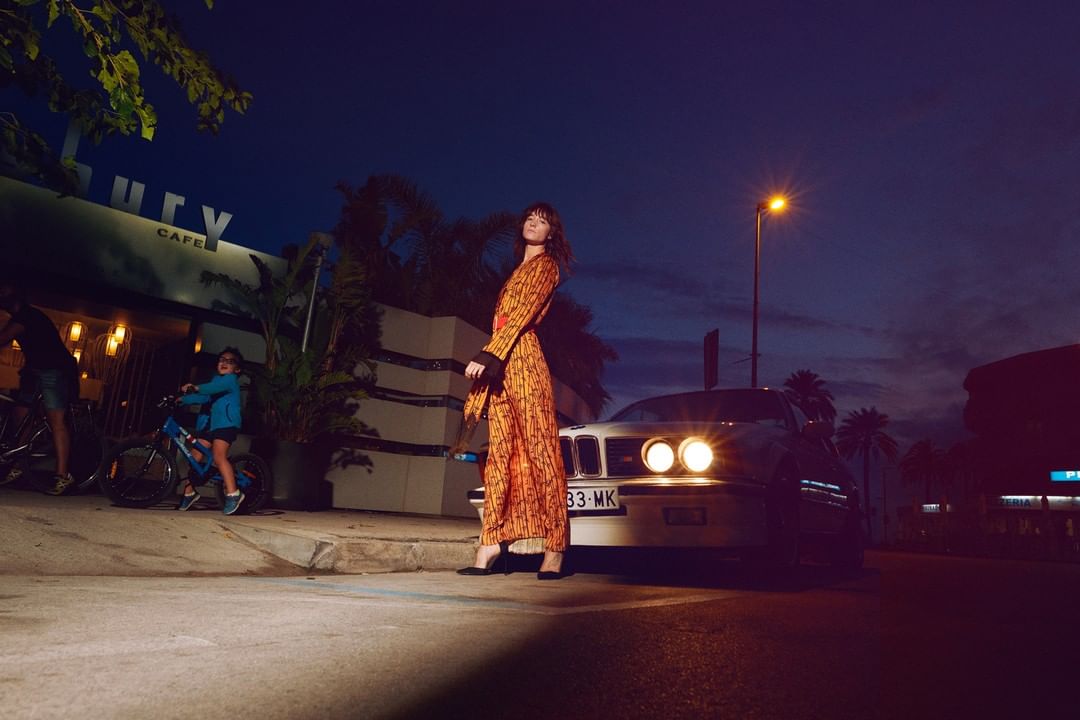 Woman poses in patterned orange suit beside vintage car and cafe at night with other people, lit by car headlights and streetlights.