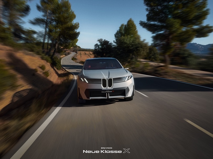 Silver BMW VISION Neue Klasse X driving on an asphalt road through a rural landscape on a sunny day with trees and mountains in the background.