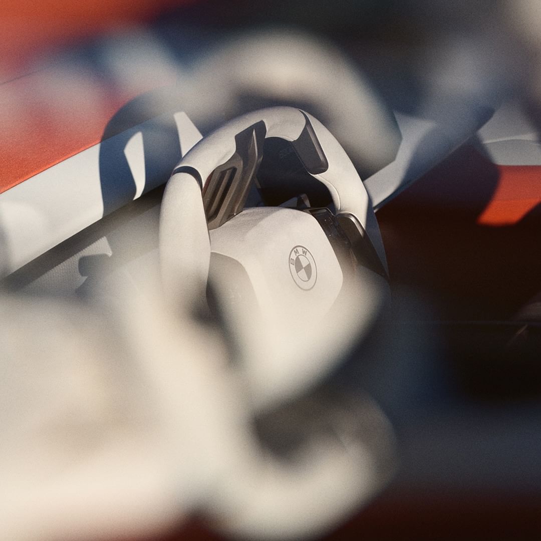 Close-up of a car interior features a white steering wheel with the BMW logo, contrasted against a red dashboard in diffused light.