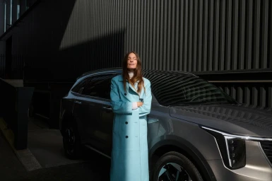 Woman in a long light blue trench coat standing with arms crossed in front of a modern grey SUV outdoors in New York
