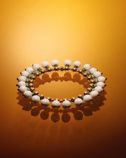 A beaded necklace with white, gold, pink, and green beads arranged in a circular pattern on a gradient yellow background.
