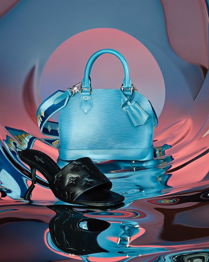 Fashion still life featuring a light blue Louis Vuitton Alma handbag with shoulder strap and a black heeled slide, on a reflective surface.