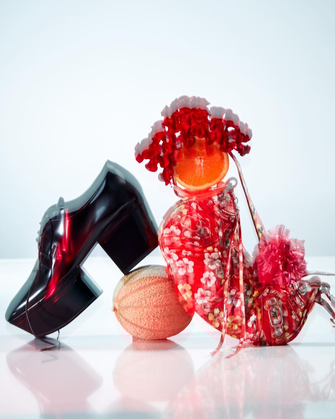 Still life of a high heel shoe, a melon, and a wig made from an orange slice and decorative accents on a white reflective surface.