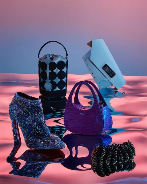 Fashion still life of designer handbags, shoes, and a bracelet on a reflective surface with soft pink and blue lighting.