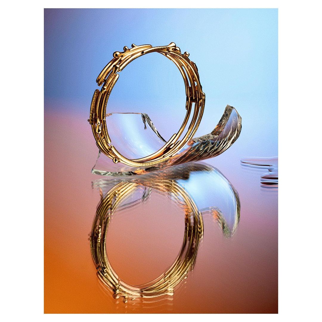 Elegant golden bracelet presented with glass shards and water droplets against a gradient blue and orange background, high fashion jewelry.