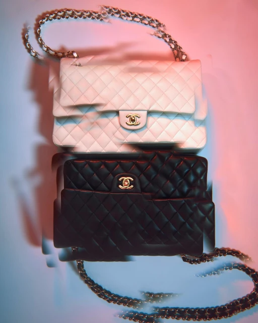 Stack of two Chanel quilted flap bags, one in white and one in black, with chain straps, lit with pink and blue studio lighting.